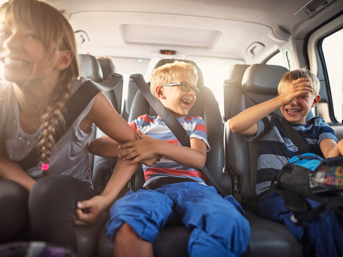 Day Trips from Nashville: Three smiling children sitting in the back seat of a car, wearing seatbelts. Two boys are laughing, while a girl with a braid grins widely. They appear to be having fun on a road trip.