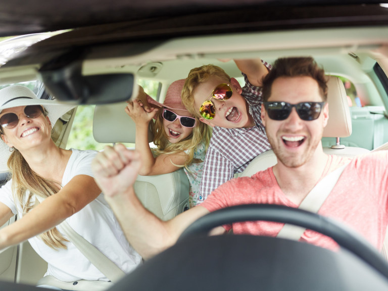 Day trips from Nashville: Happy family of four smiling and cheering inside a car, wearing sunglasses and hats, enjoying a fun road trip together.