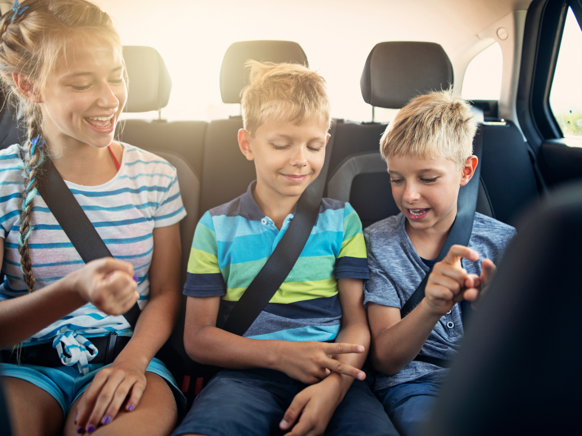 Day trips from Austin: Three kids sitting in the back seat of a car, wearing seat belts, smiling, and playing a game together during a road trip.