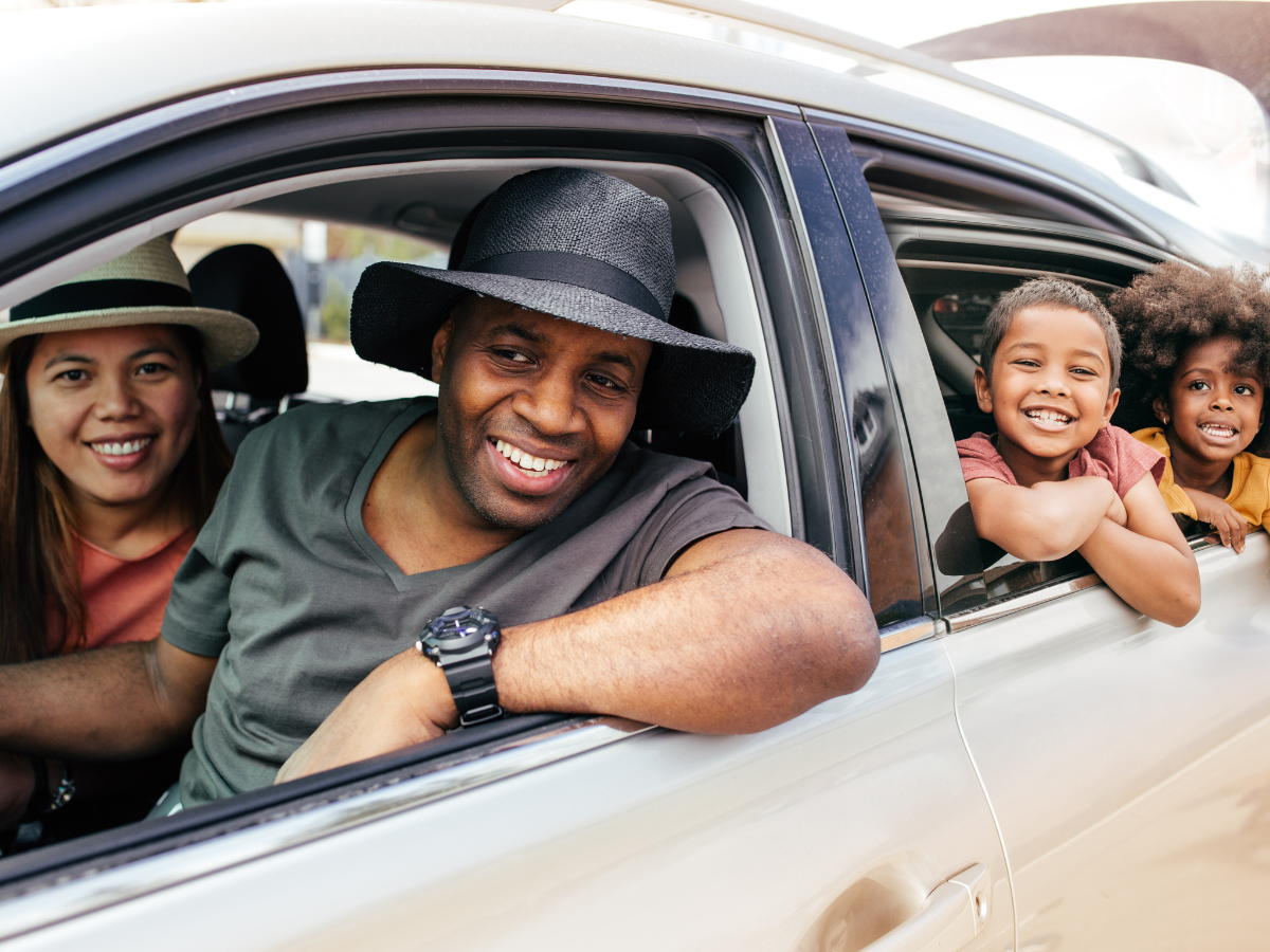 Day trips from Charlotte: A cheerful family in a car, smiling through open windows, ready for a day trip. The parents are in the front seats wearing hats, and two children are leaning out from the back seat, all looking excited.