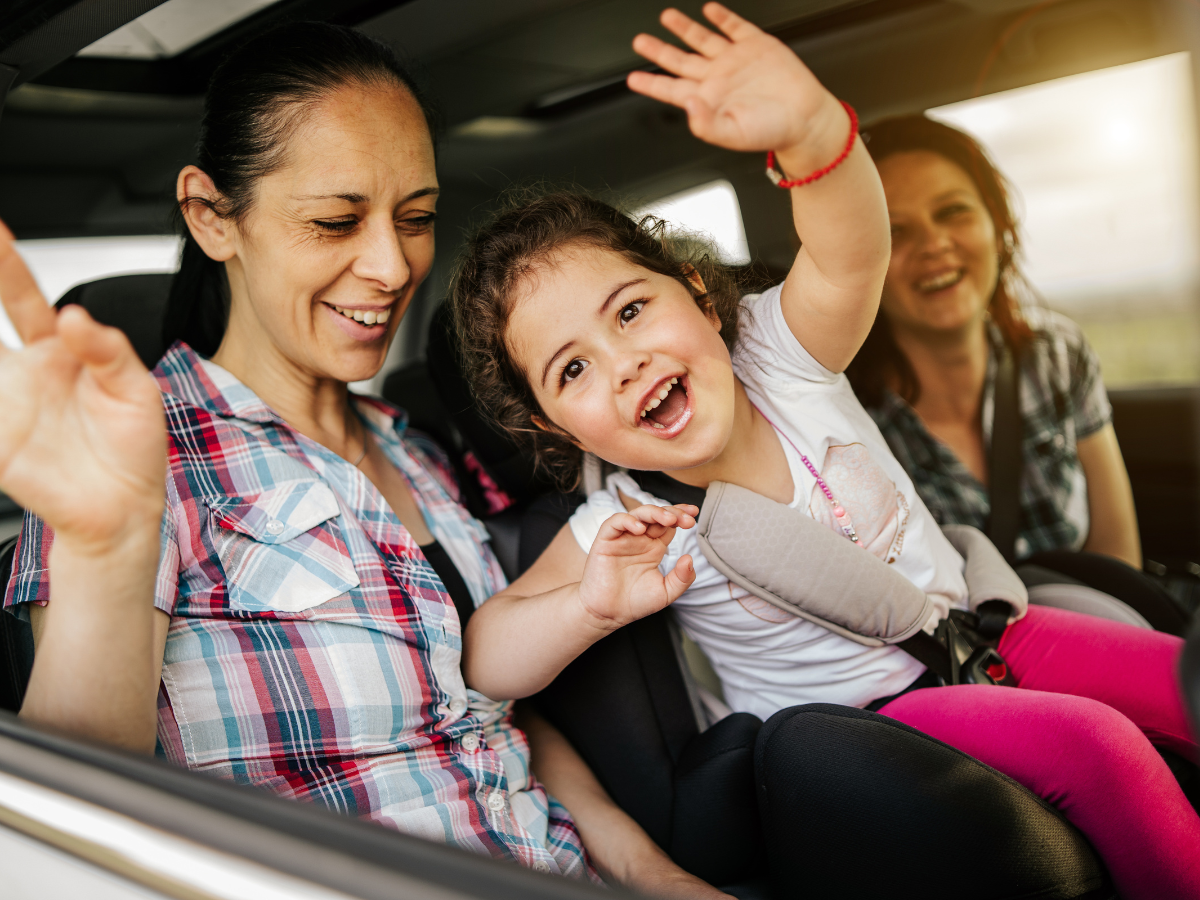 Day trip from Charlotte: A joyful child in a car seat, waving excitedly with two smiling adults sitting beside her. The family appears to be enjoying a road trip together.