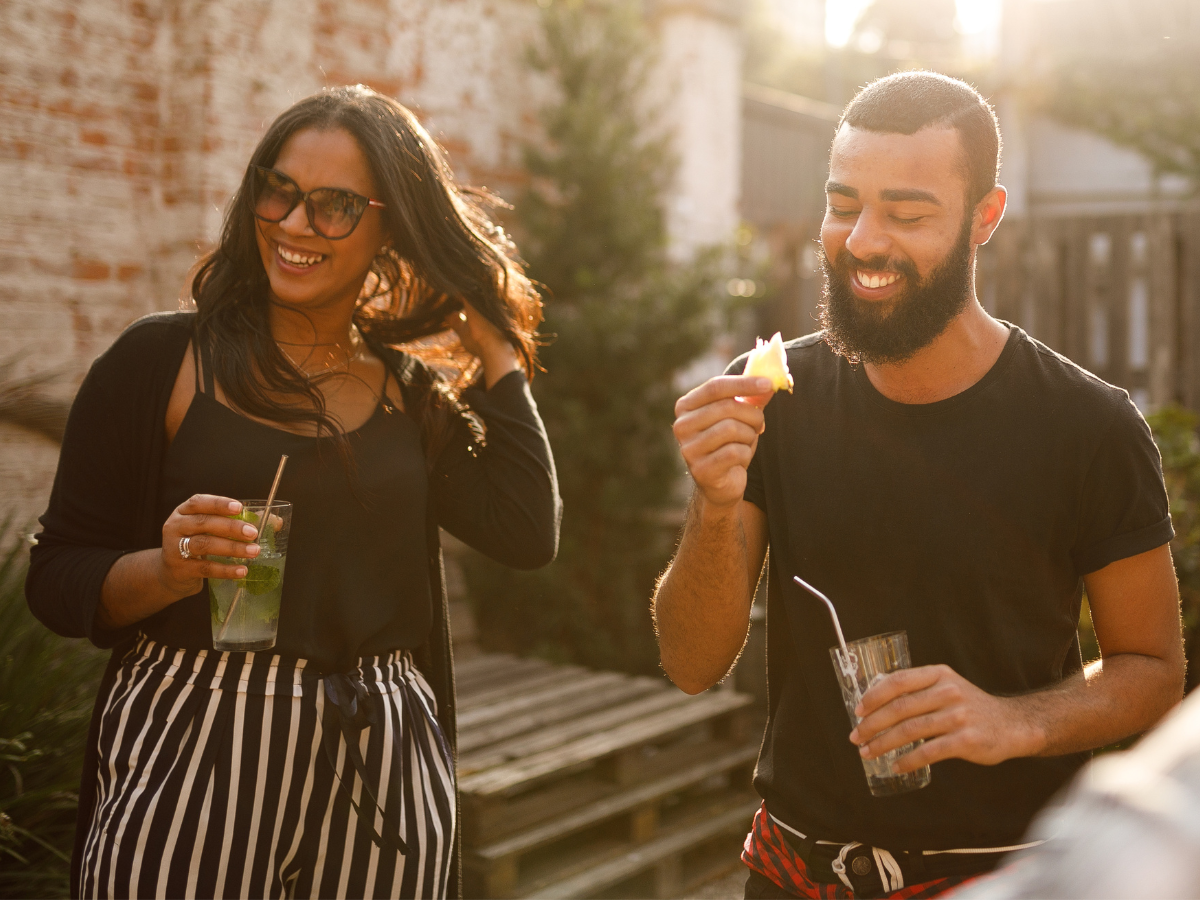 date night ideas in Charlotte: A smiling couple enjoying a casual outdoor date. The woman holds a cocktail while adjusting her hair, and the man laughs while holding a drink in one hand and his garnish in the other.