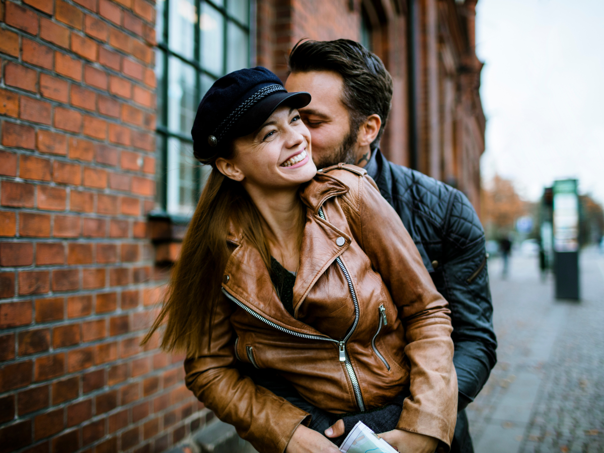 date night ideas in Washington, DC: A happy couple embracing outdoors on a city street. The woman is wearing a brown leather jacket and a hat, while the man hugs her from behind, both smiling and enjoying a romantic moment