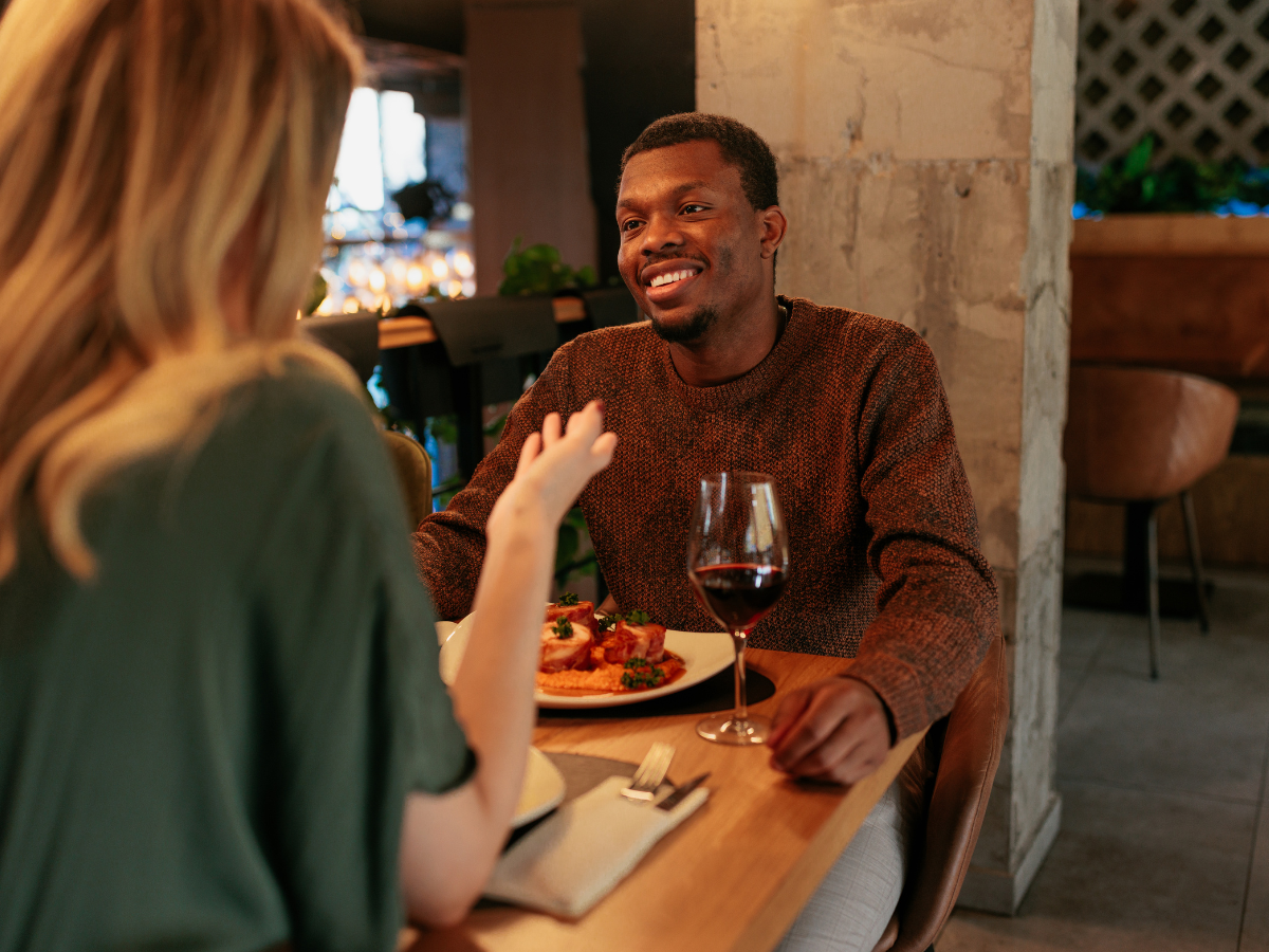 date night ideas in Washington, DC: A couple enjoying a dinner date at a cozy restaurant. The man is smiling warmly across the table, with a glass of red wine in front of him, while the woman gestures as she speaks