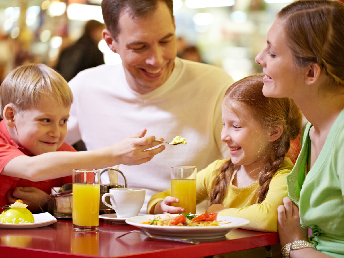 kids eat free restaurants: A cheerful family enjoying a meal at a restaurant. The parents smile while the young boy playfully feeds his sister, creating a warm and happy atmosphere