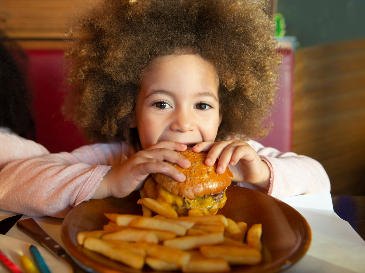 kids eat free restaurants: A young child with curly hair enjoying a burger at a restaurant, with a plate of fries in front of them. The child is smiling and holding the burger with both hands