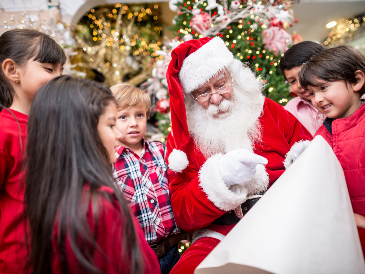 Photos with Santa in Tampa: Santa Claus surrounded by smiling children, holding a large scroll in a festive setting with a decorated Christmas tree in the background