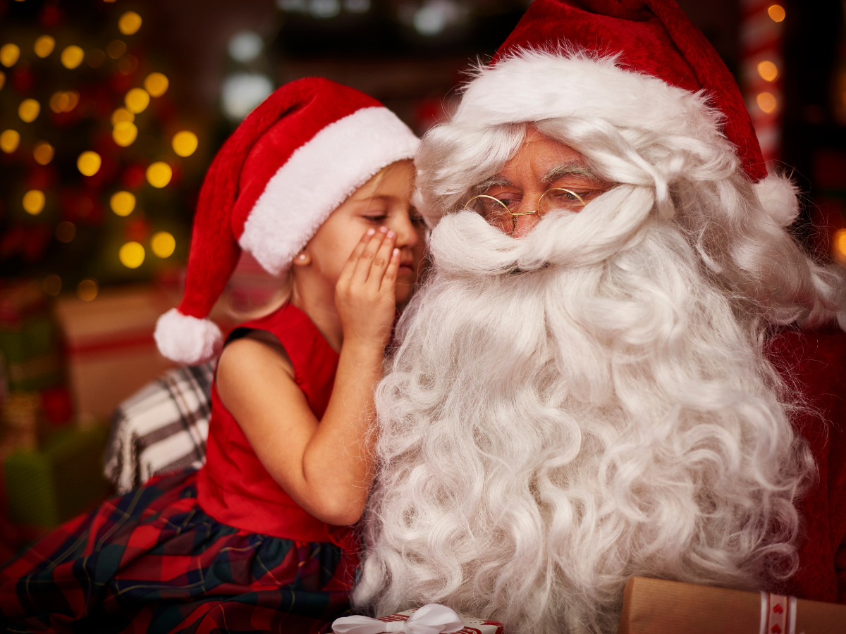 Photos with Santa in Tampa: A young child in a Santa hat whispering to Santa Claus, surrounded by festive holiday decorations and warm lighting