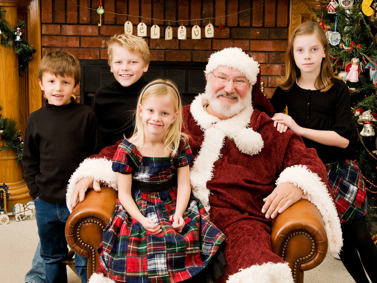 Photos with Santa in Orlando: Santa Claus posing with four smiling children in holiday attire in a cozy, festive room decorated with a Christmas tree and 'Believe' sign