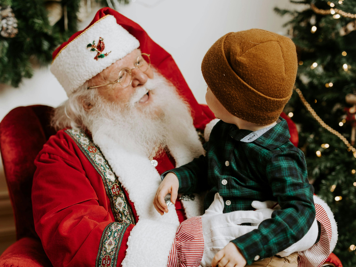 Photos with Santa in Denver: A young child sitting on Santa Claus's lap, looking up at him with a Christmas tree in the background, creating a warm holiday moment