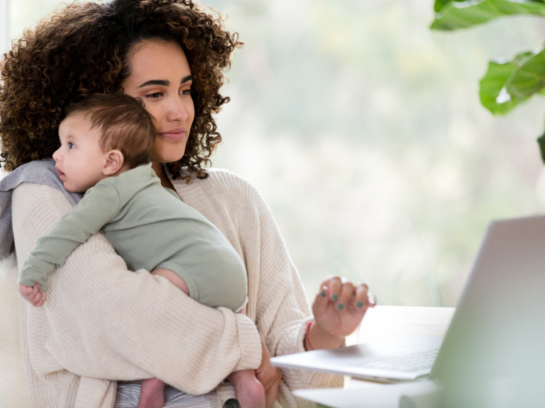 What to Know About Background Checks: A mother holding her baby looks at her laptop, appearing focused and thoughtful. The natural light and cozy setting create a warm, family-oriented atmosphere