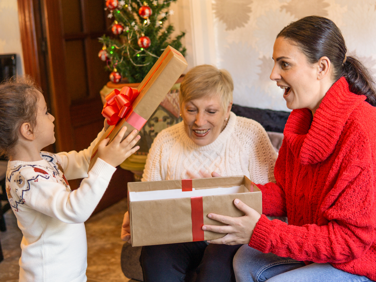 Holiday Gift Guide for Mom: A young child hands a wrapped gift to her smiling mother, while a grandmother watches joyfully beside them, seated near a decorated Christmas tree