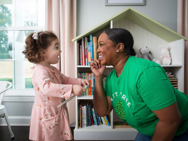 Preparing for a Babysitting Job: A babysitter wearing a green SitterTree shirt smiles and interacts playfully with a young child holding a book, standing in a cozy playroom with a bookshelf in the background