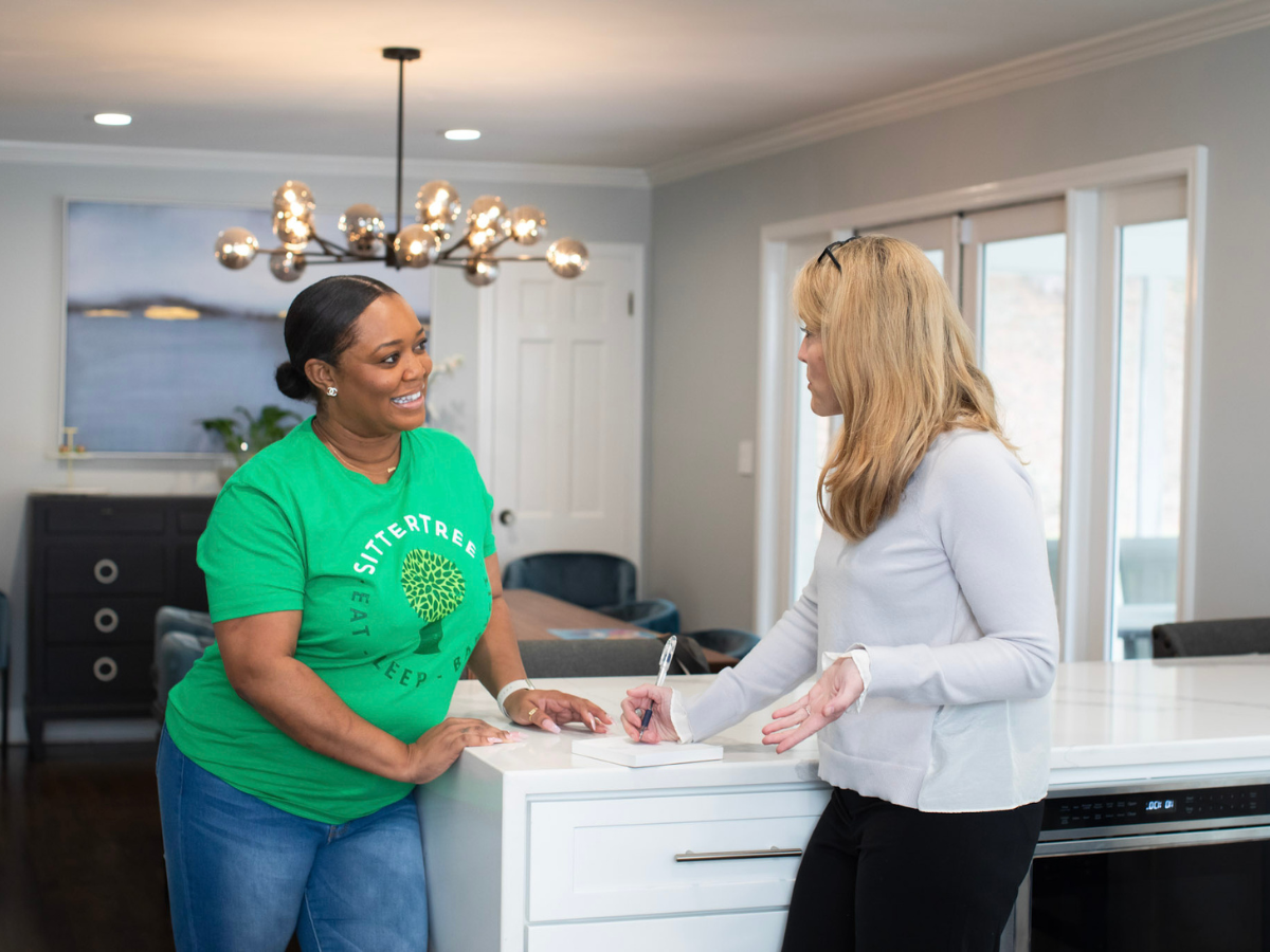 Preparing for a Babysitting Job: A babysitter wearing a green SitterTree shirt stands in a kitchen, smiling and having a conversation with a parent holding a pen and notebook, discussing job details