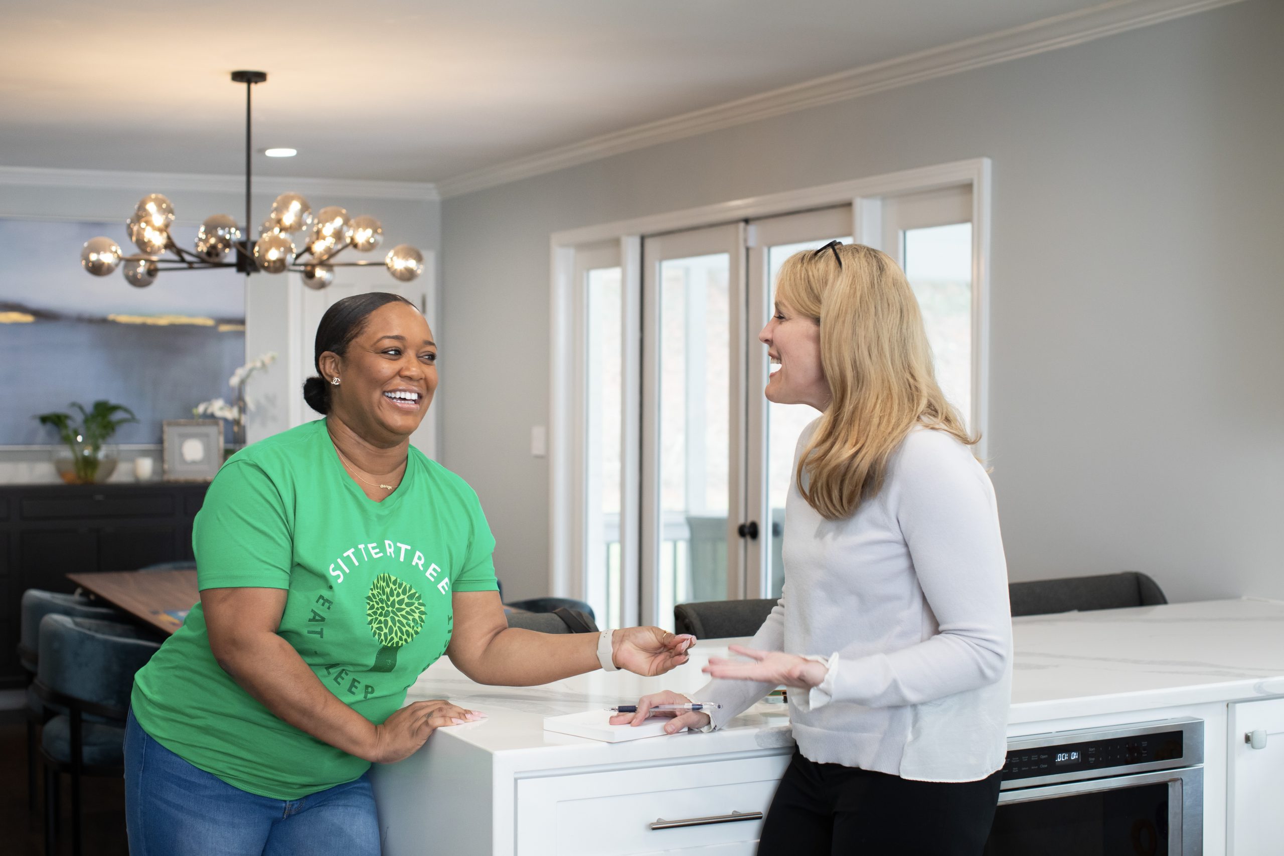 Tips for screening your babysitter: A SitterTree sitter in a green T-shirt shares a warm, friendly conversation with a parent in a bright, modern kitchen. Both are smiling and appear engaged, creating a welcoming and relaxed atmosphere