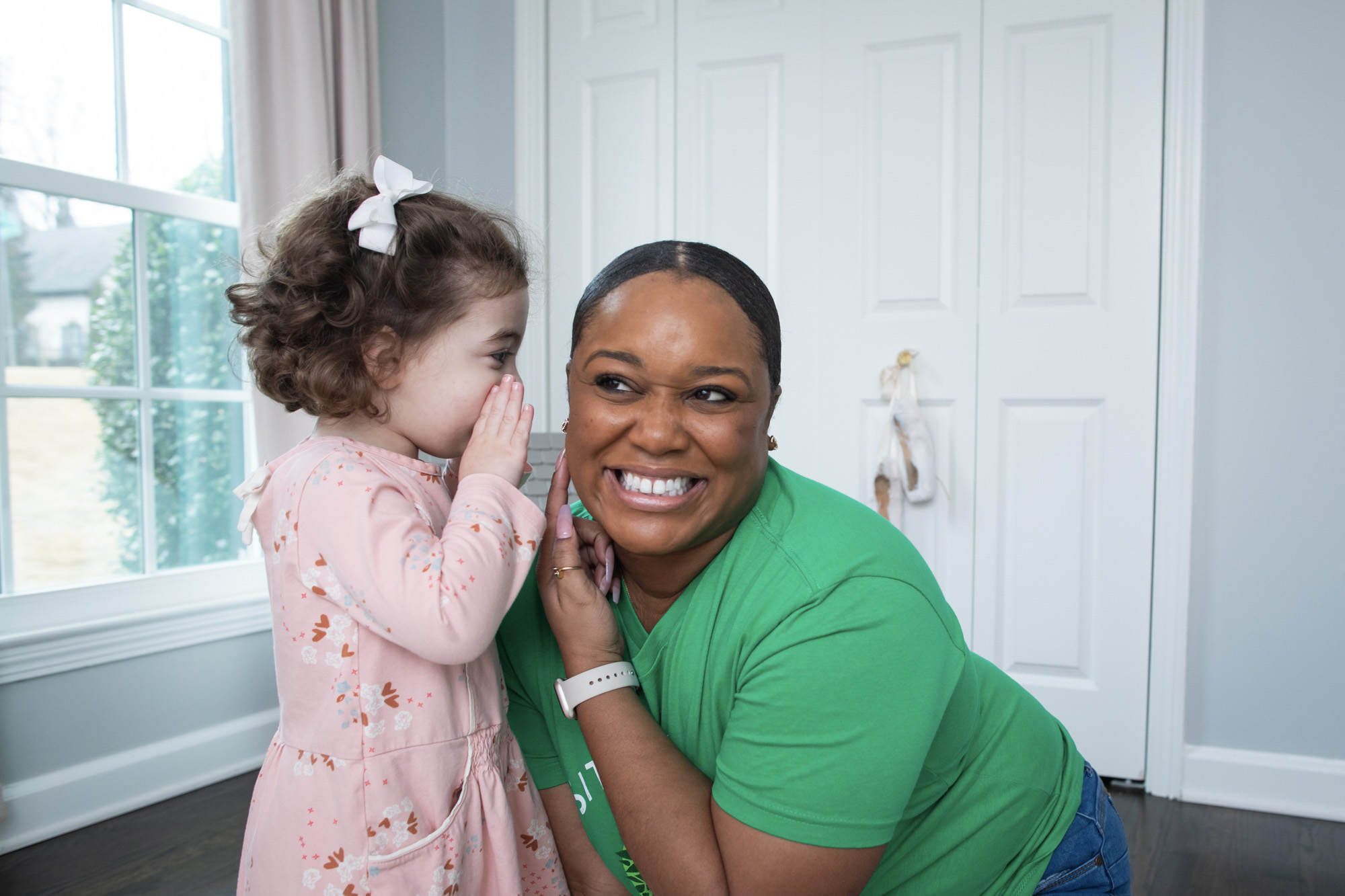 How much to pay a babysitter per hour: A young girl whispers into the ear of a smiling babysitter wearing a green SitterTree t-shirt, sharing a playful and joyful moment indoors