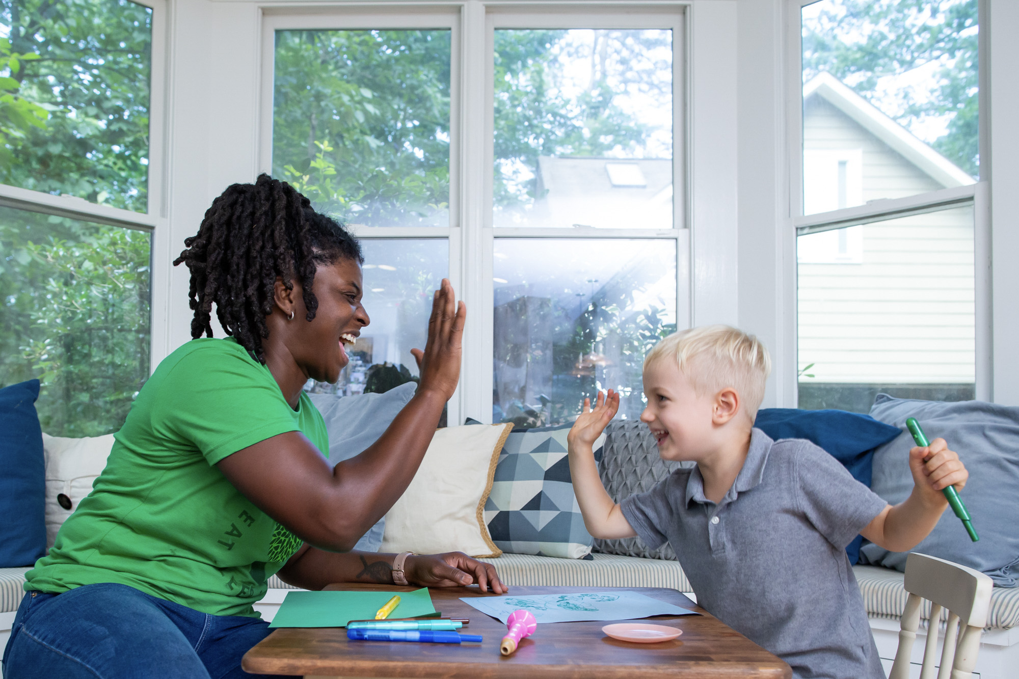 Questions to ask a nanny in an interview: A SitterTree caregiver and a young boy share a joyful high-five during an art activity at a small table. The caregiver smiles warmly as the boy holds a marker, creating a fun and engaging moment in a bright, cozy room