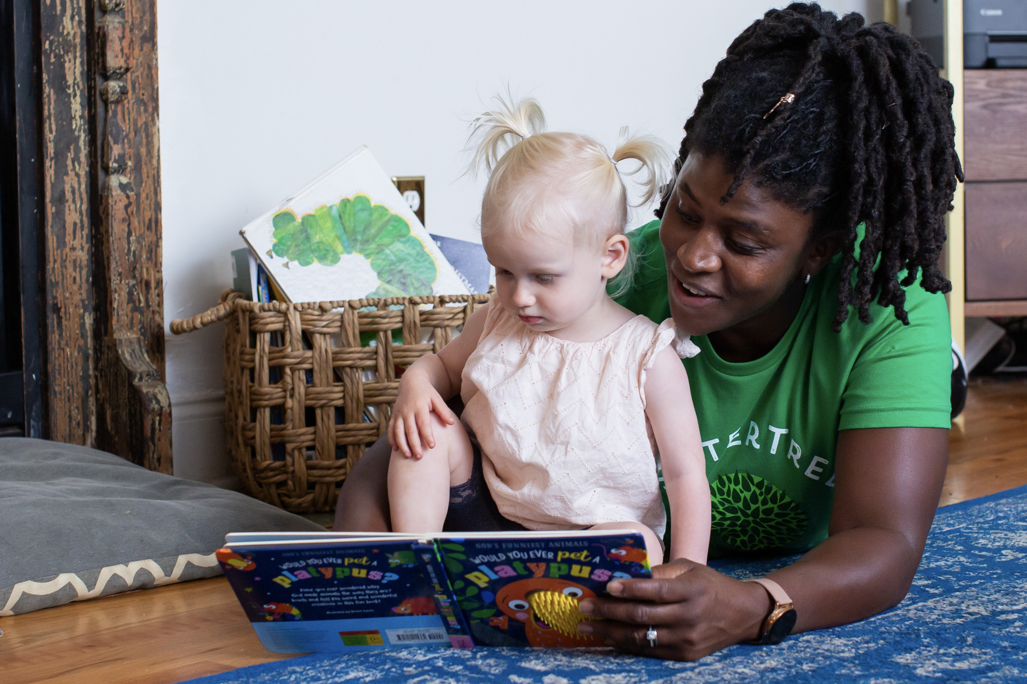 How much does a babysitter cost per month: A babysitter wearing a green SitterTree t-shirt reads a colorful children's book to a toddler with blonde pigtails, creating a cozy and engaging moment