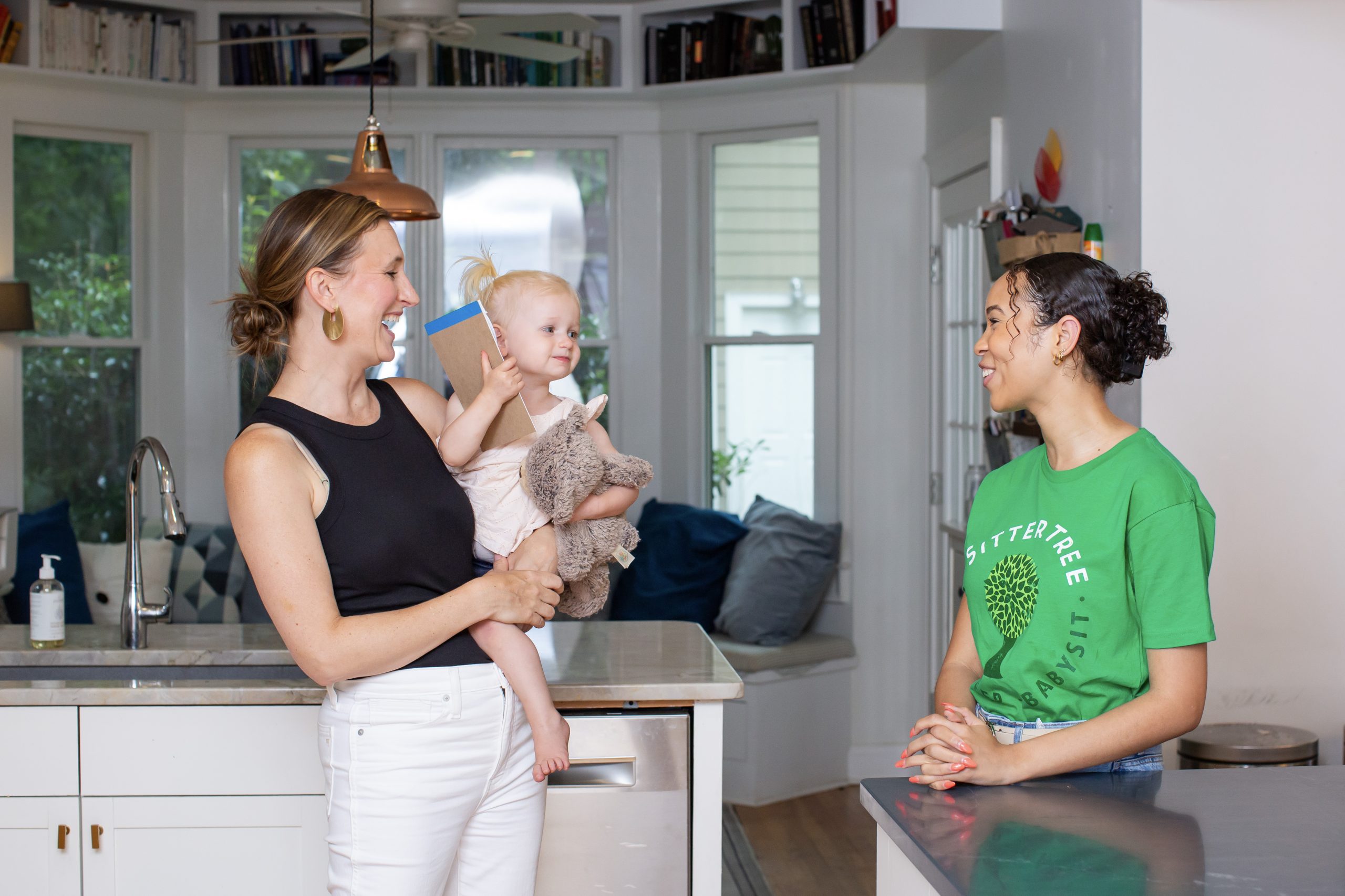 Tips for Screening Your Babysitter: A smiling SitterTree sitter chats with a mom holding her young child in a cozy kitchen. The child holds a stuffed animal and a notebook, creating a warm, friendly moment between the sitter and family.