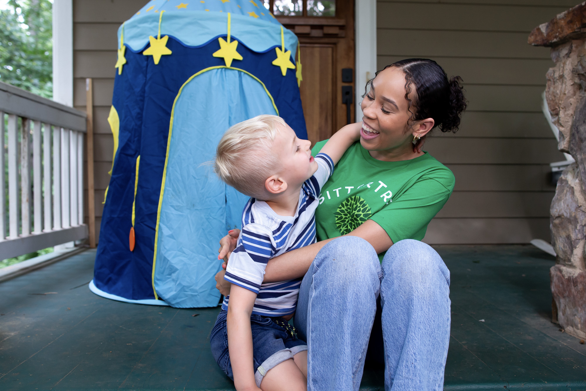 Questions to ask a babysitter: A SitterTree caregiver sitting on a porch, smiling and interacting with a young boy in a striped shirt. They share a joyful moment as the boy leans in with his arm around her, with a play tent visible in the background