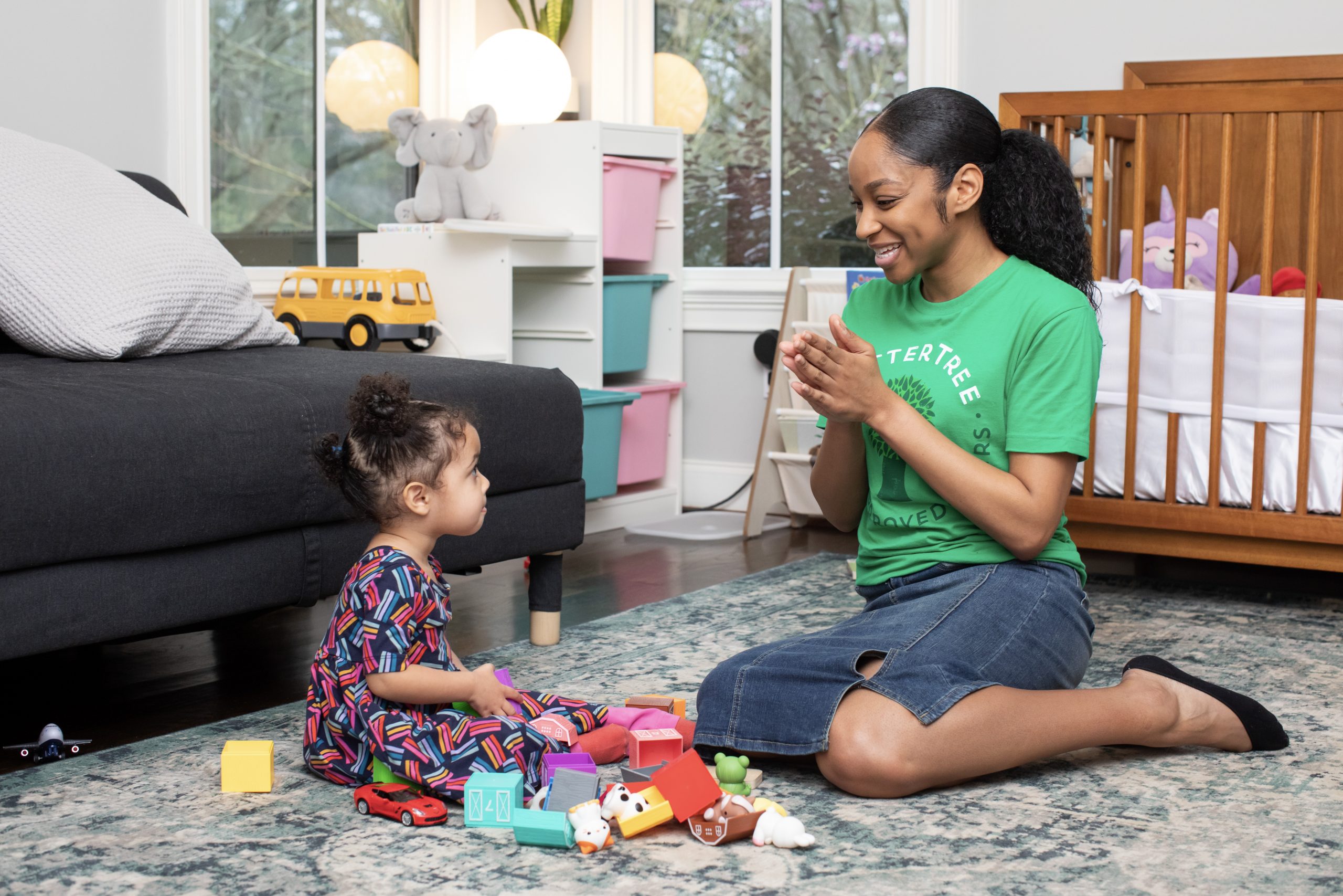 How to Prepare for a New Babysitter: A SitterTree sitter claps and smiles while engaging with a young child who is sitting on the floor surrounded by toys. They are in a cozy, well-organized playroom, creating a warm and playful atmosphere