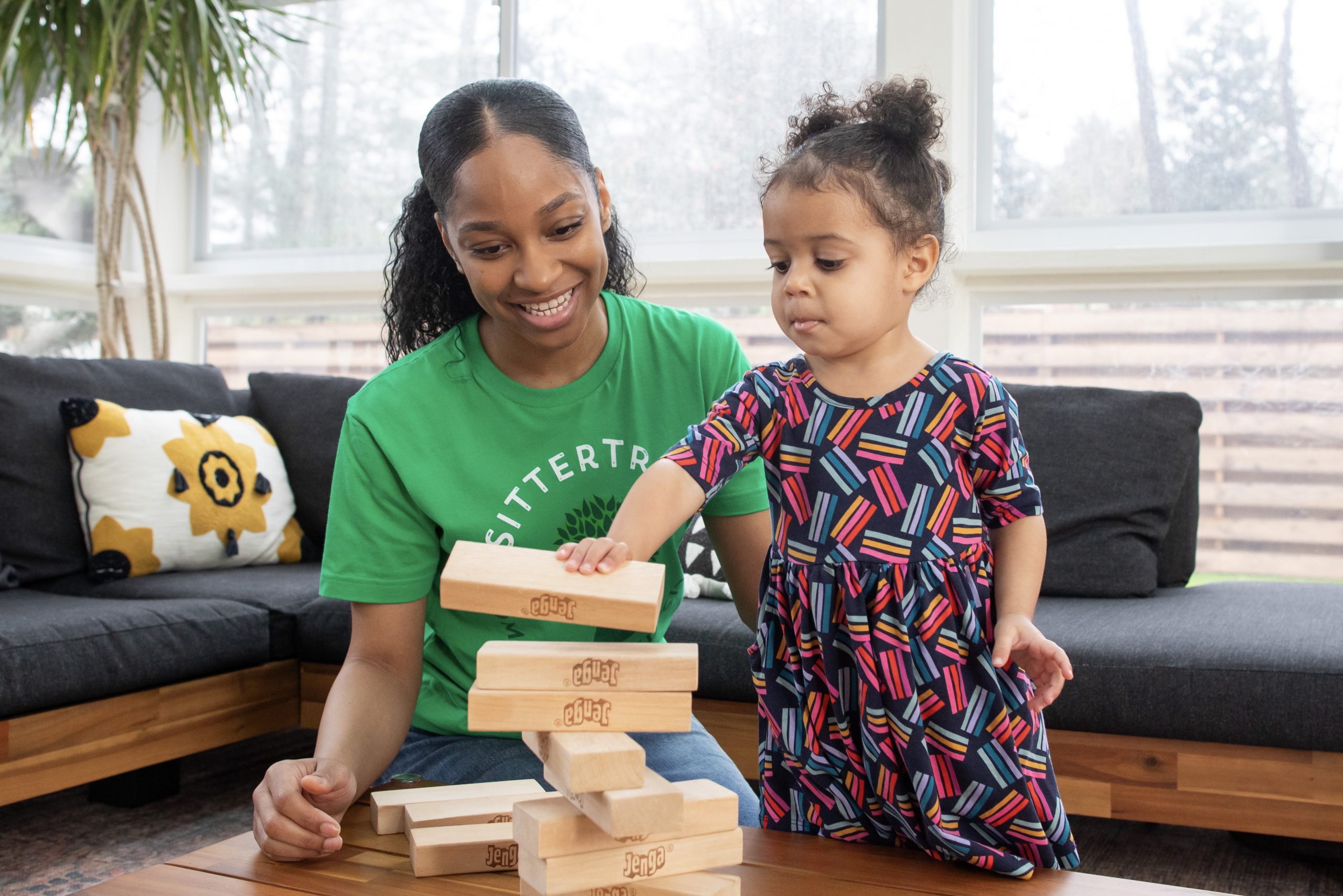 Questions to ask a babysitter: A SitterTree caregiver and a young girl playing a game of Jenga together in a bright living room. The caregiver smiles as the child carefully removes a block, creating a fun and engaging moment