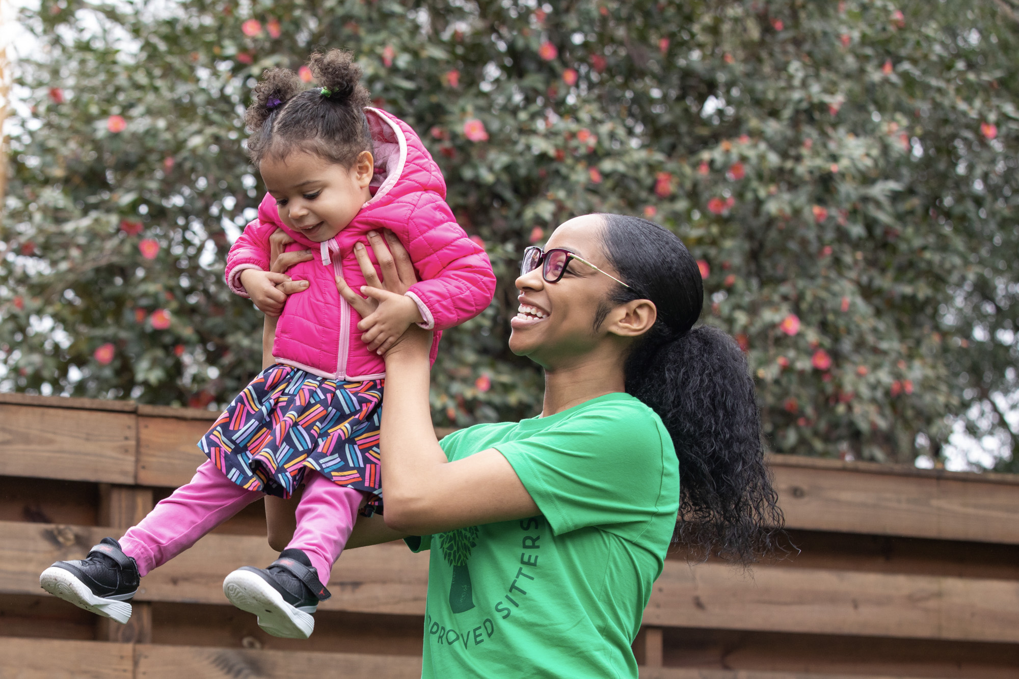 Choosing between a babysitter and a nanny: A SitterTree caregiver smiling and playing outdoors with a young girl wearing a bright pink jacket. The caregiver is lifting the child joyfully, creating a fun, interactive moment