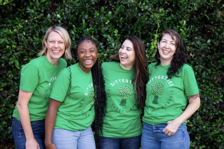 How much to charge for babysitting jobs: Four women wearing green SitterTree t-shirts smiling and laughing together in front of a lush green hedge, exuding a sense of camaraderie and joy