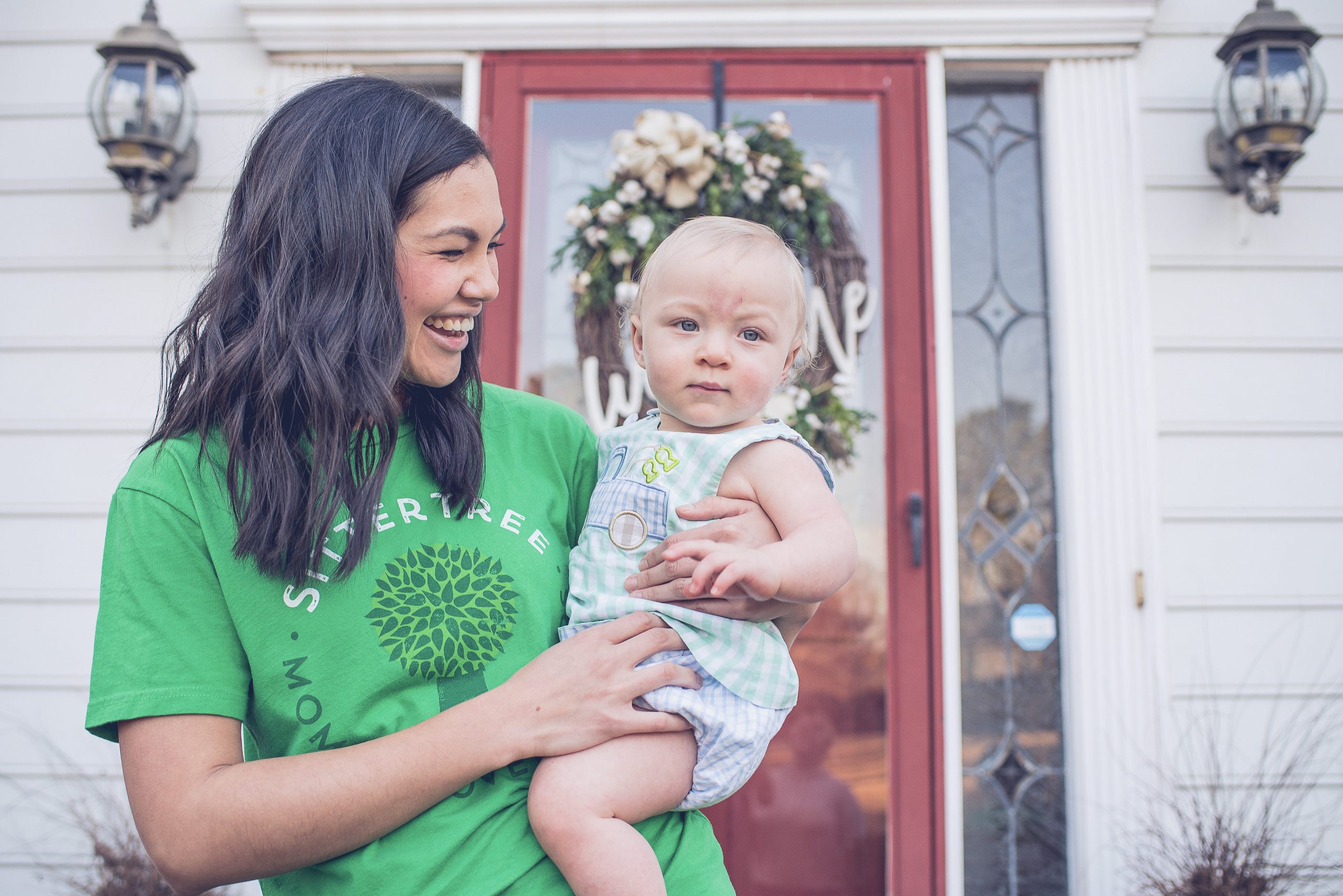 How much to charge for babysitting jobs: A smiling SitterTree babysitter wearing a green branded shirt holds a baby outside a home with a festive wreath on the red door