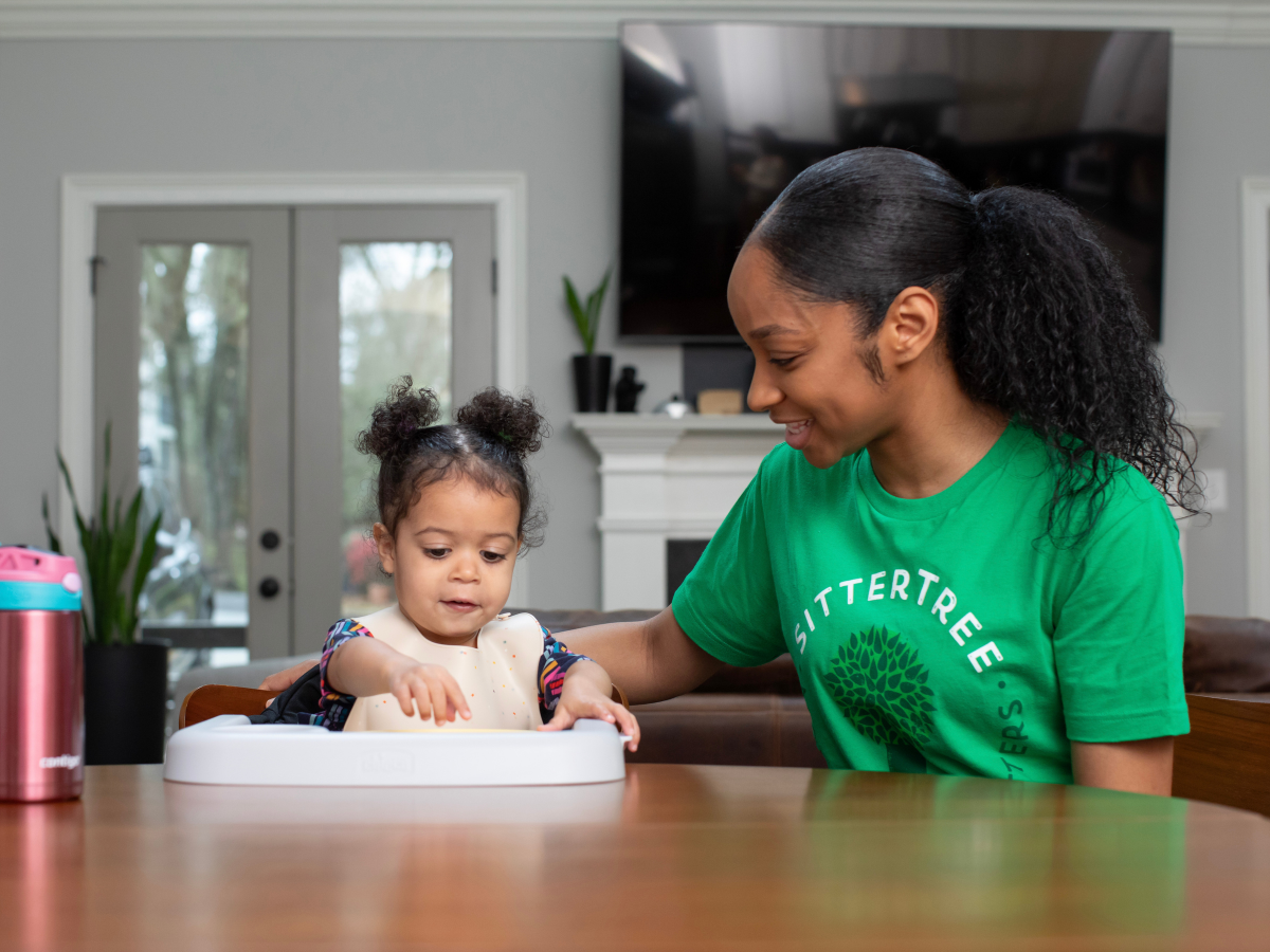 Babysitter vs. Nanny: A SitterTree sitter wearing a green SitterTree T-shirt smiles while engaging with a young toddler in a highchair at a dining table, creating a warm and caring environment in a modern home.