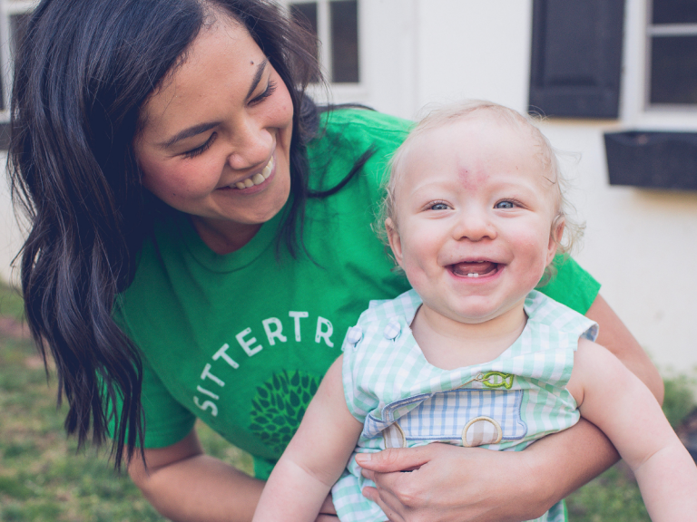 Safety tips for sitters: A smiling SitterTree sitter wearing a green SitterTree T-shirt holding a happy baby outdoors, capturing a joyful and caring moment