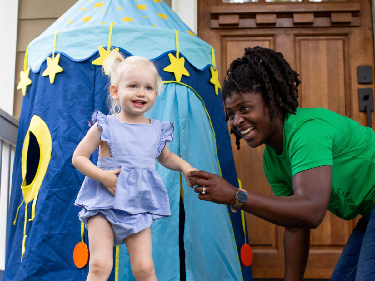 Safety tips for sitters: A smiling SitterTree sitter wearing a green SitterTree T-shirt playing with a happy toddler near a colorful play tent on a porch, capturing a fun and caring moment