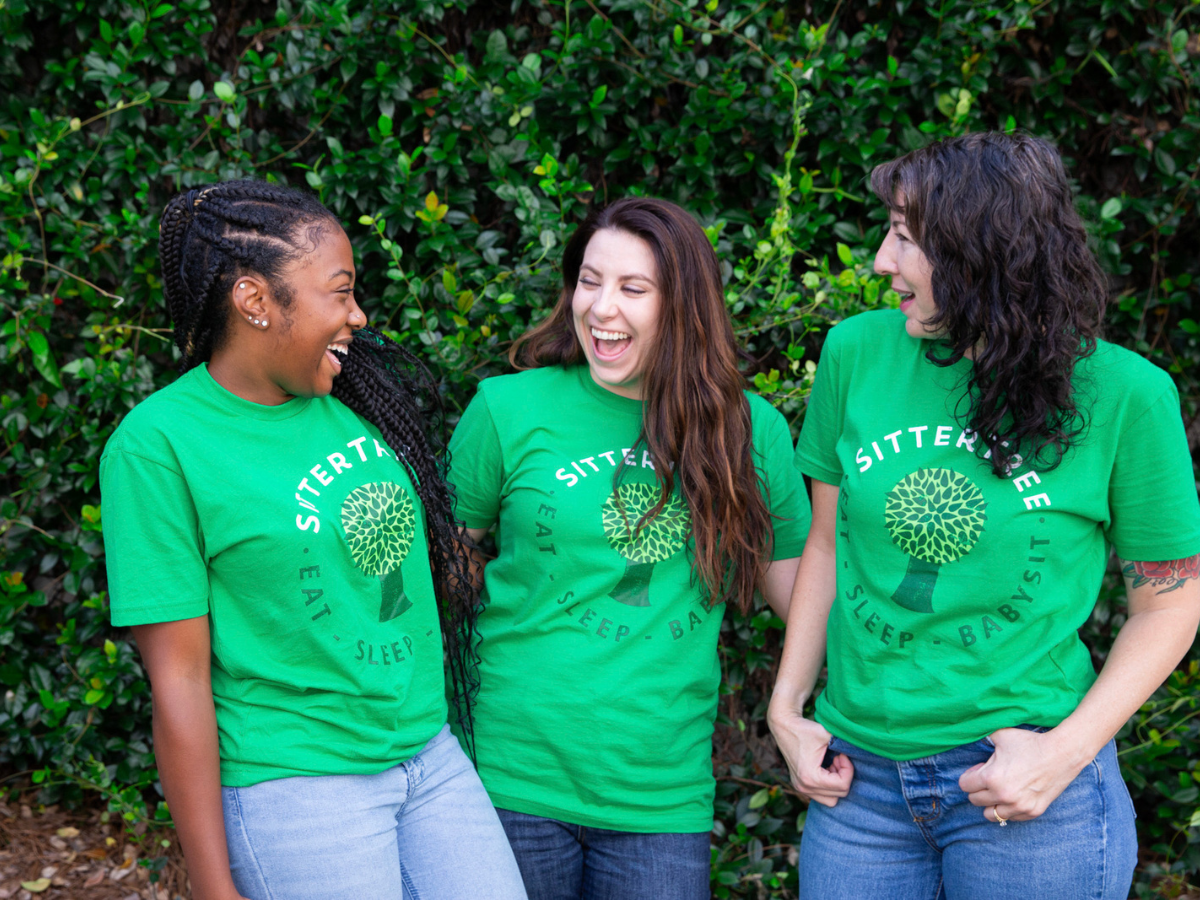 How to gain babysitting experience: Three smiling SitterTree sitters wearing green SitterTree T-shirts laughing together outdoors in front of lush greenery, highlighting camaraderie and joy