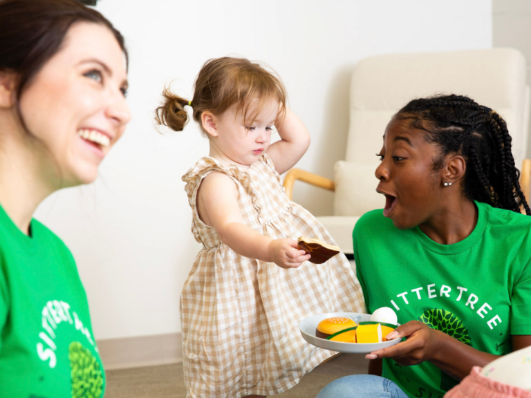 background checks for babysitters: A smiling SitterTree sitter wearing a green SitterTree T-shirt playfully interacting with a toddler holding a toy, creating a fun and engaging moment