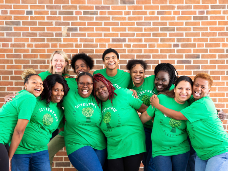 Babysitter vs. Nanny: A group of diverse sitters wearing green SitterTree T-shirts, smiling and embracing each other in front of a brick wall, showcasing a sense of community and teamwork.