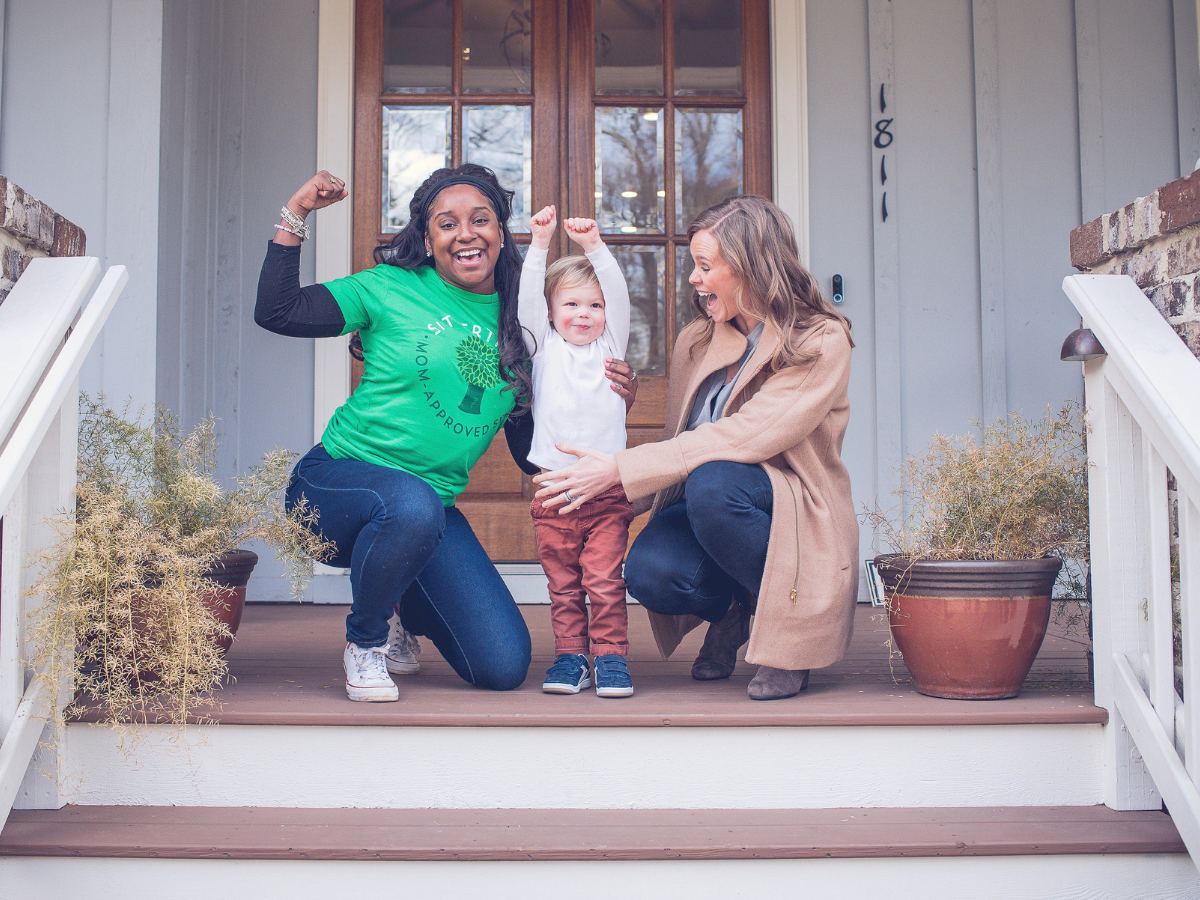 background checks for babysitters: "Mom-Approved Sitter" T-shirt flexing her arms in a playful pose with a toddler and a parent on the front porch of a home, capturing a joyful and trusting moment