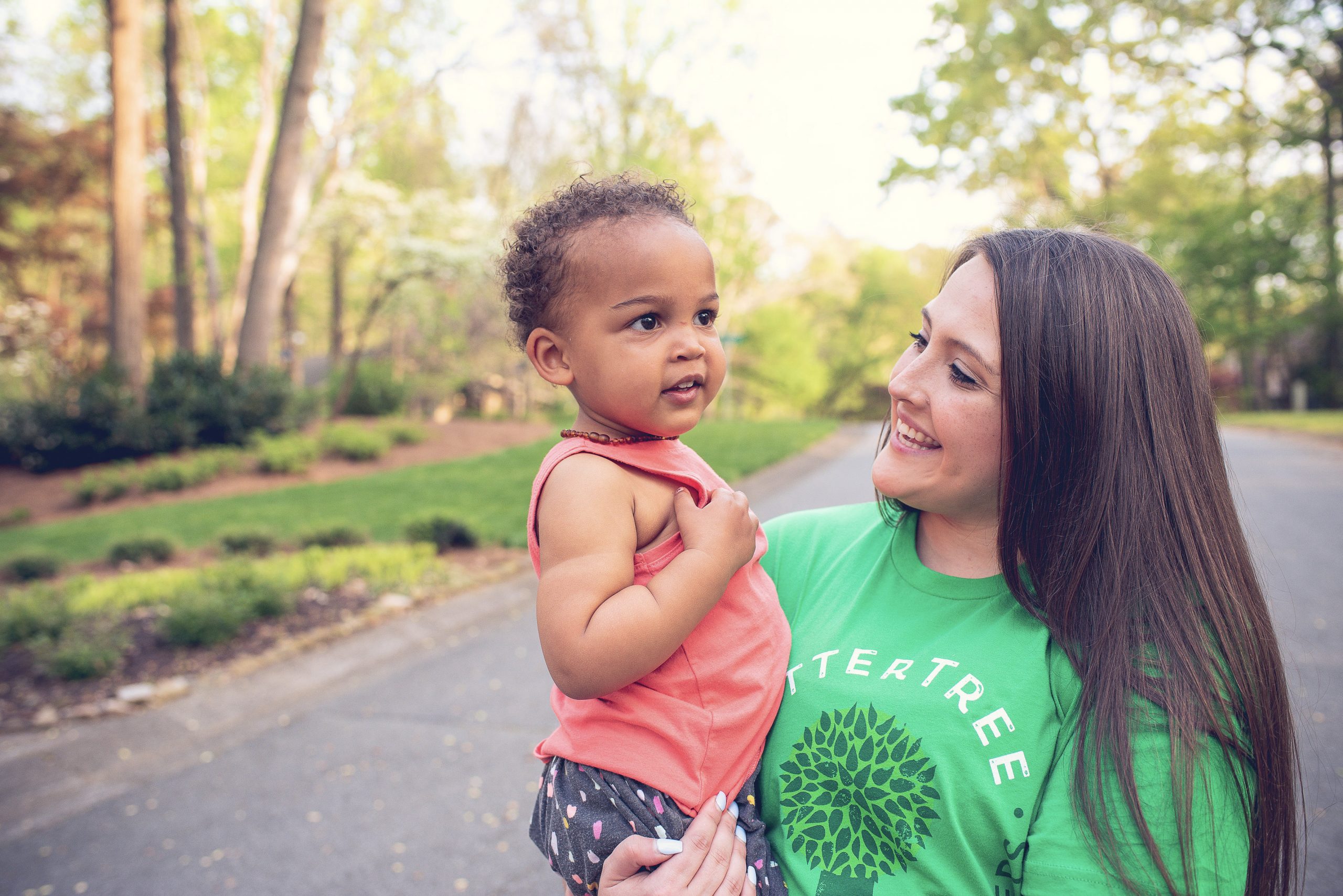 A smiling woman wearing a green SitterTree T-shirt holds a young child outdoors on a sunny day, with a tree-lined path in the background: First-time babysitter tips