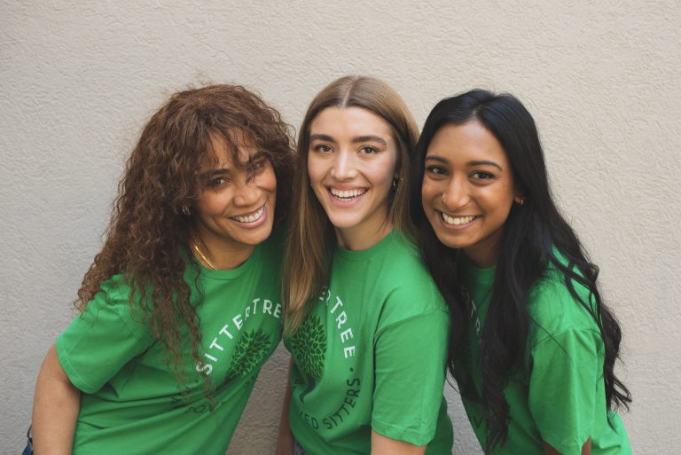 Three smiling women wearing green SitterTree T-shirts, standing closely together against a light-colored wall: How to transition from babysitter to nanny