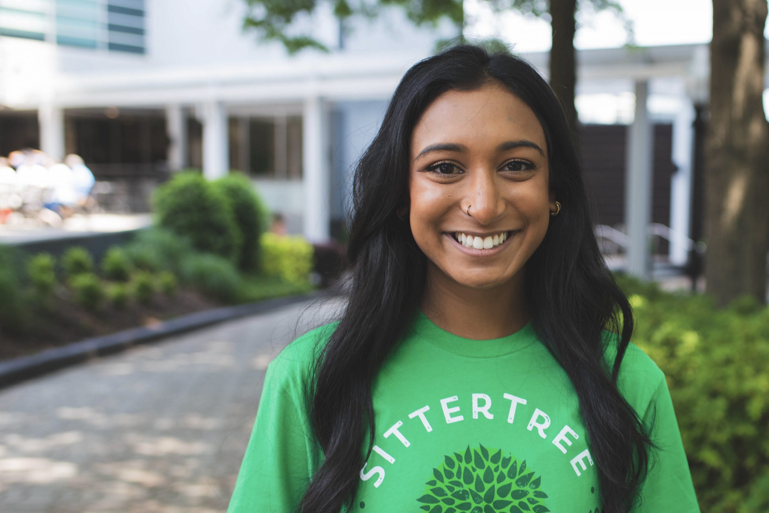 A smiling woman wearing a green SitterTree T-shirt stands outdoors in front of a landscaped area with trees and a modern building in the background: How to transition from babysitter to nanny