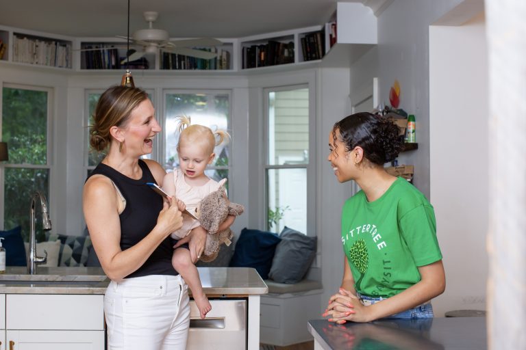 A smiling mother holding her toddler, who is clutching a stuffed toy, chats with a babysitter wearing a green SitterTree t-shirt in a bright and cozy kitchen: How to ace a babysitting interview