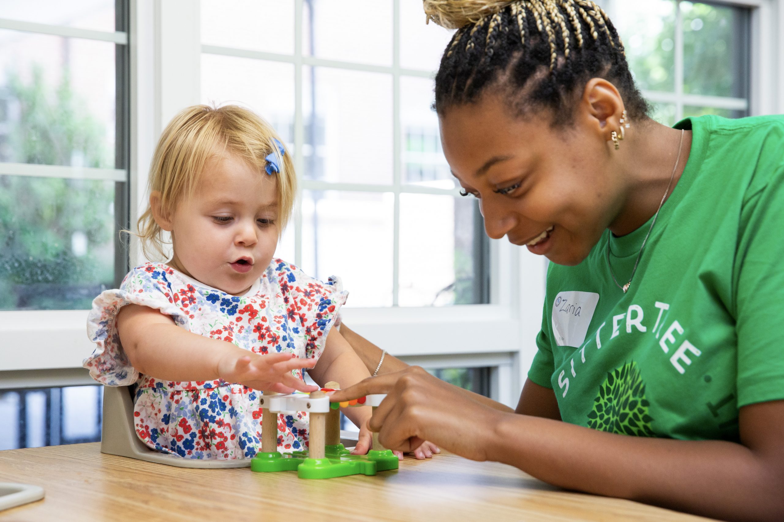 A young woman wearing a green SitterTree t-shirt interacts with a toddler playing with a toy at a wooden table in a brightly lit room: Best babysitting apps and websites