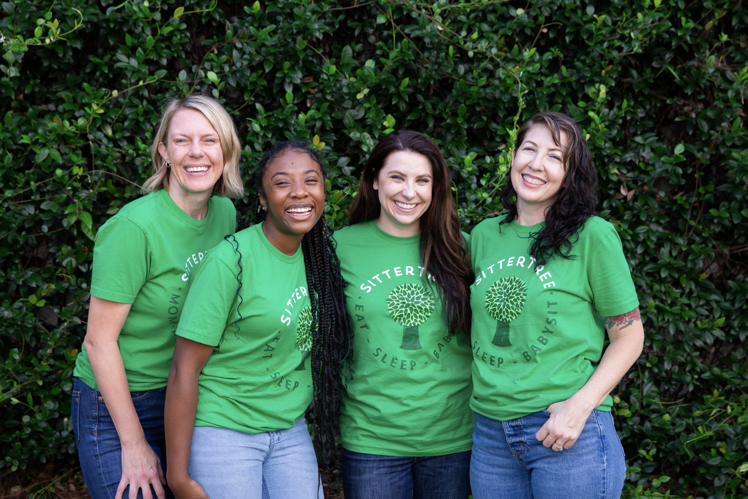 A group of four smiling babysitters wearing green SitterTree t-shirts stand in front of a lush green hedge, showcasing camaraderie and team spirit: How to become a babysitter