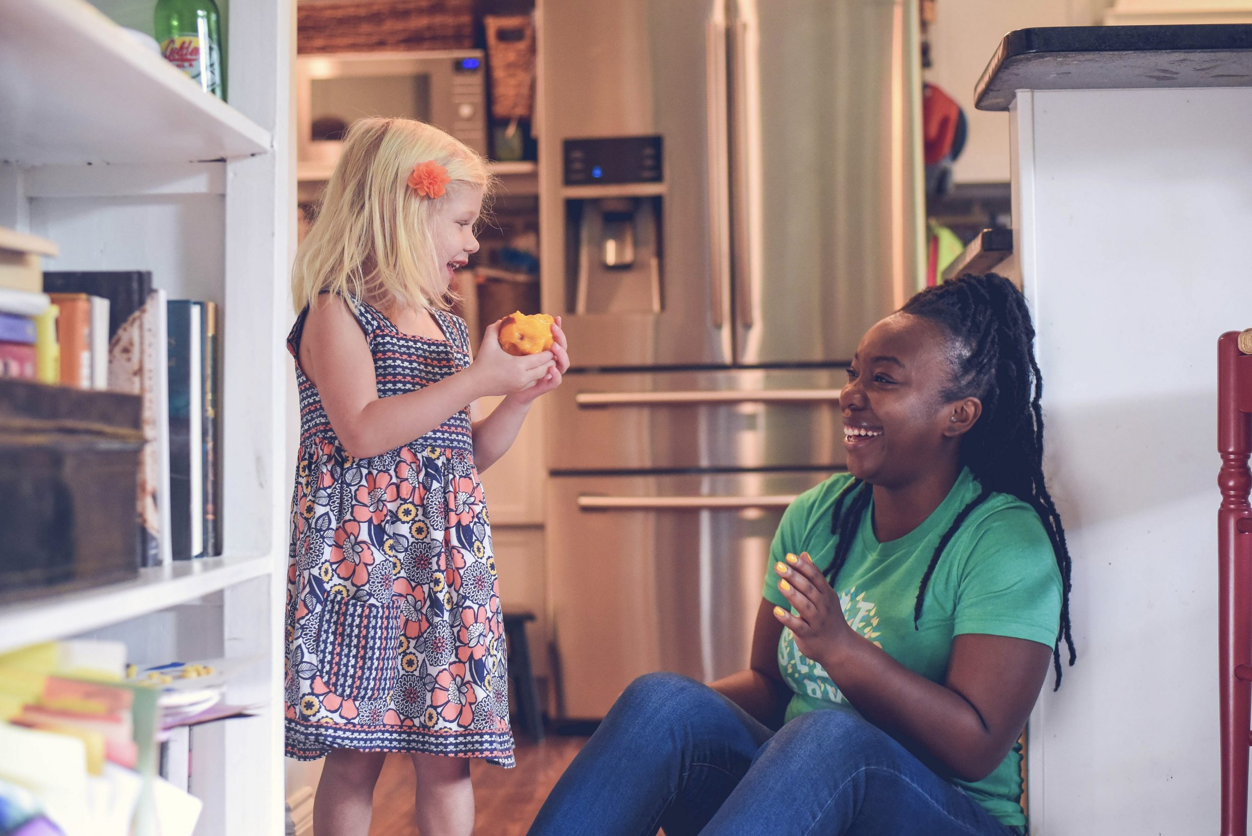 A babysitter wearing a green SitterTree t-shirt sits on the kitchen floor, smiling and engaging with a young girl holding a peach, surrounded by a cozy home setting: Babysitting kit essentials