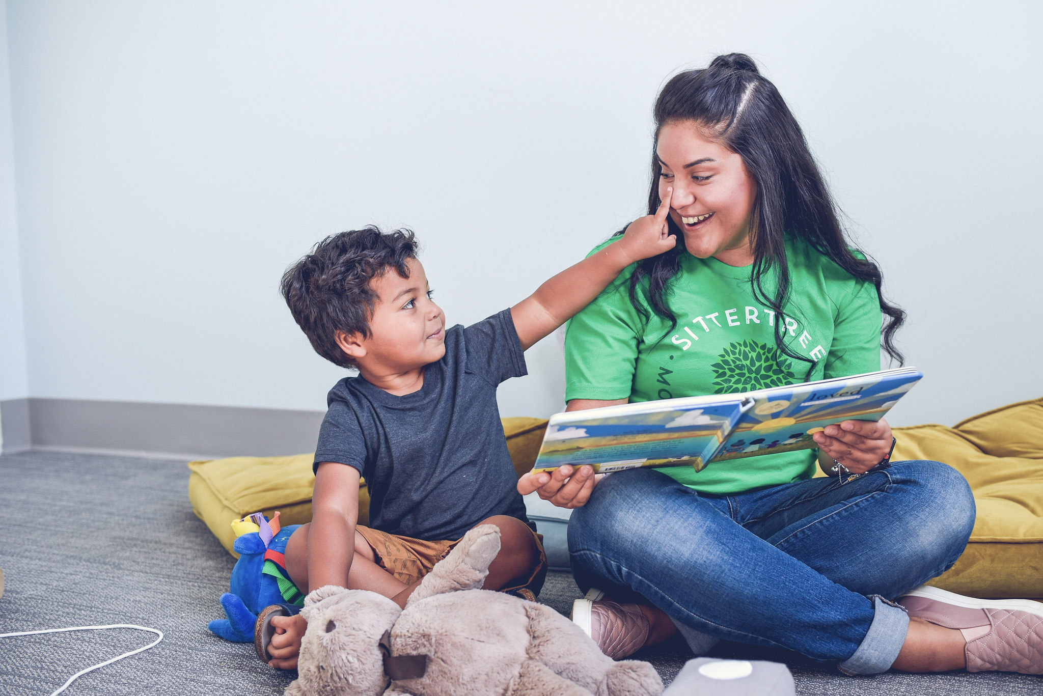 A babysitter wearing a green SitterTree t-shirt sits on the floor reading a book to a young boy, who playfully touches her face, surrounded by stuffed toys and cushions: Babysitting kit essentials