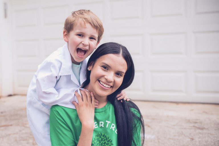 A babysitter wearing a green SitterTree t-shirt smiles as a young boy dressed as a doctor playfully leans on her shoulders outdoors: Creative play ideas for school-aged children