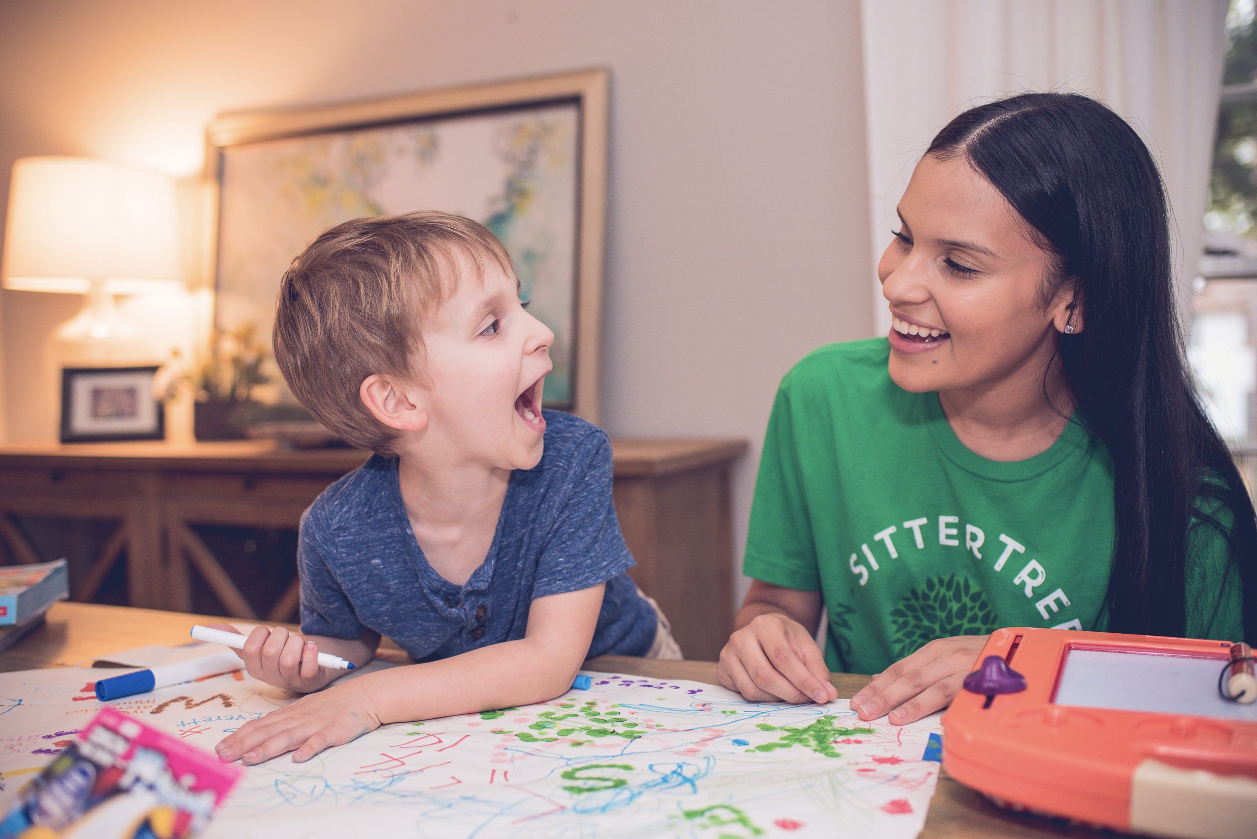 A babysitter wearing a green SitterTree t-shirt laughs with a young boy as they draw together on a colorful piece of paper in a cozy room: Creative play ideas for school-aged children