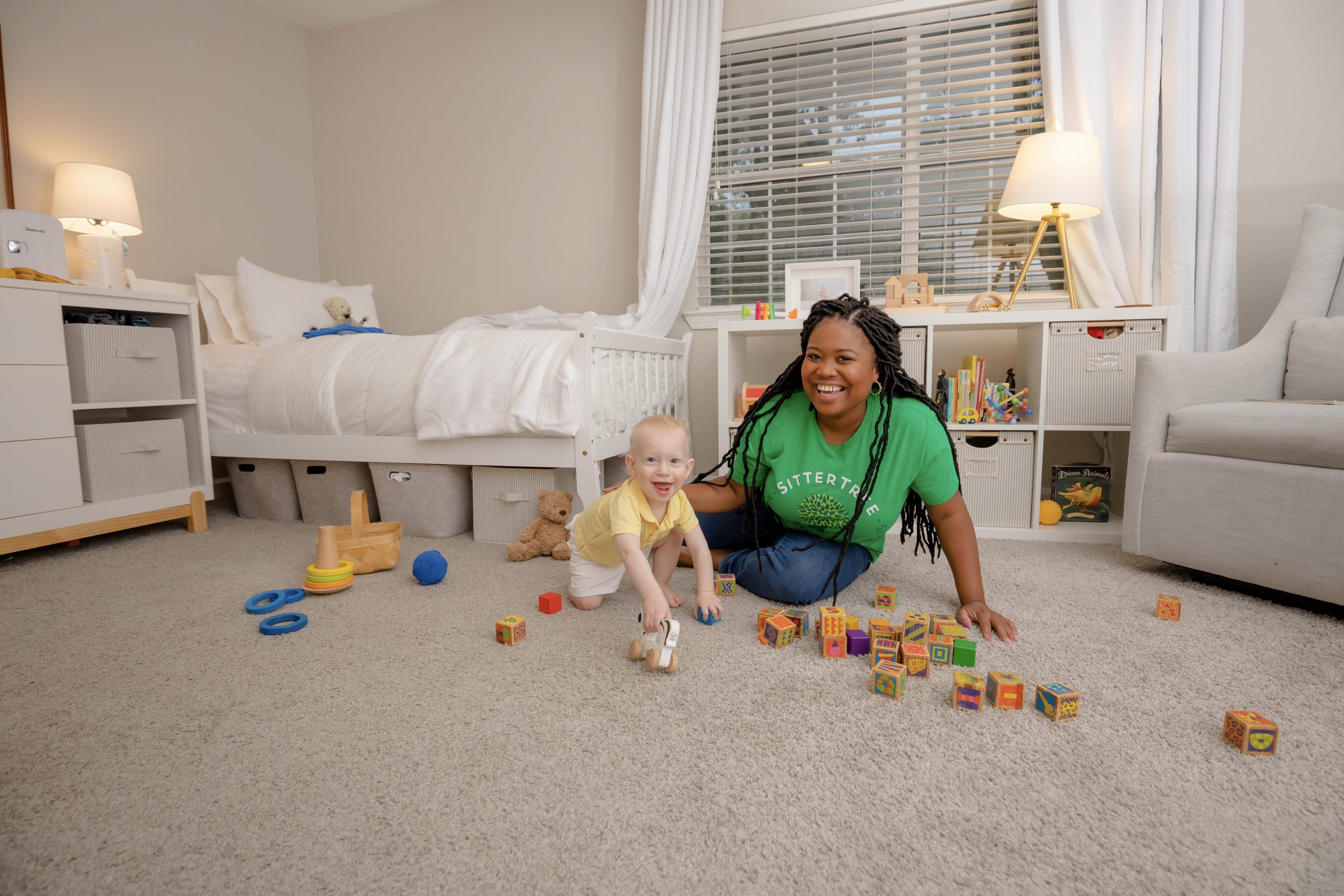 A smiling woman wearing a green SitterTree T-shirt plays on the floor with a baby surrounded by colorful toys in a cozy, well-organized bedroom: Best babysitting certification programs