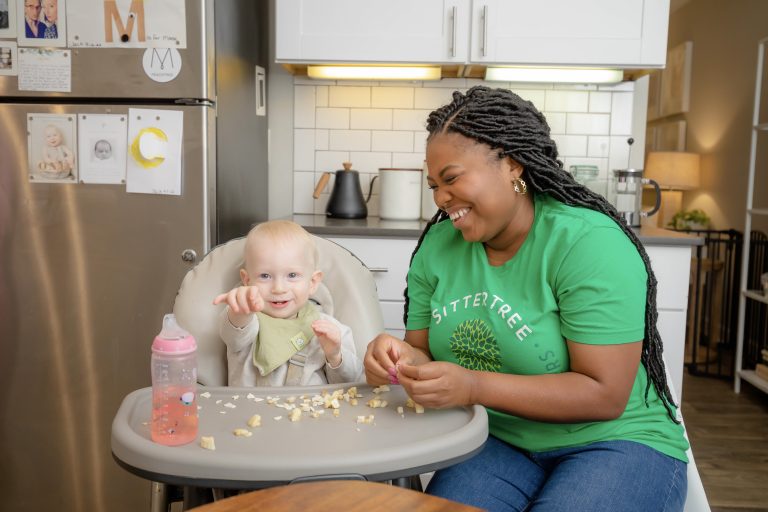 A smiling woman wearing a green SitterTree T-shirt sits next to a baby in a high chair, sharing a playful moment in a modern kitchen: Best babysitting certification programs