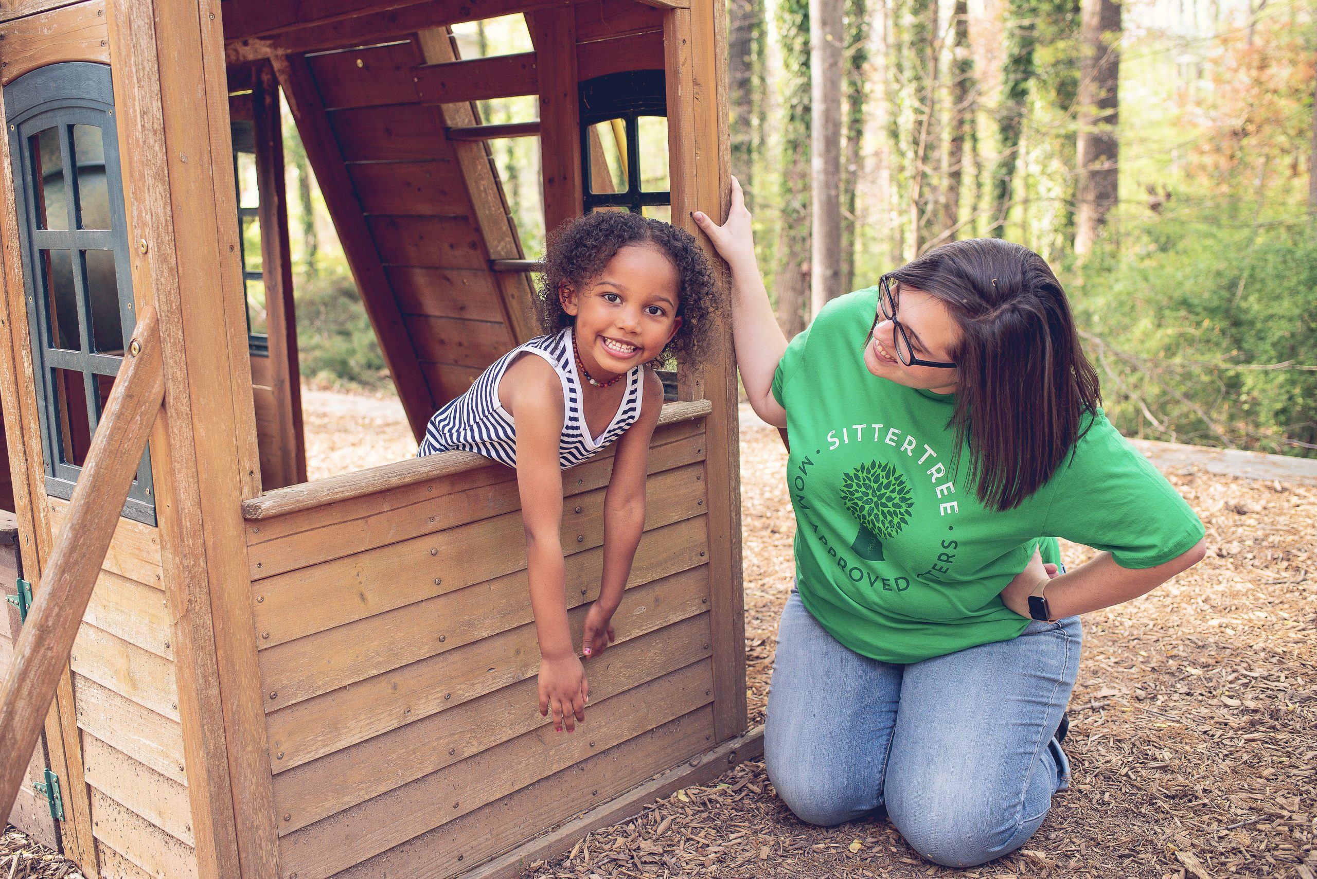 Smiling young girl leaning out of a wooden playhouse window, while a babysitter in a green SitterTree t-shirt kneels nearby, engaging with her in a playful outdoor setting surrounded by trees: how to maintain open communication with your babysitter