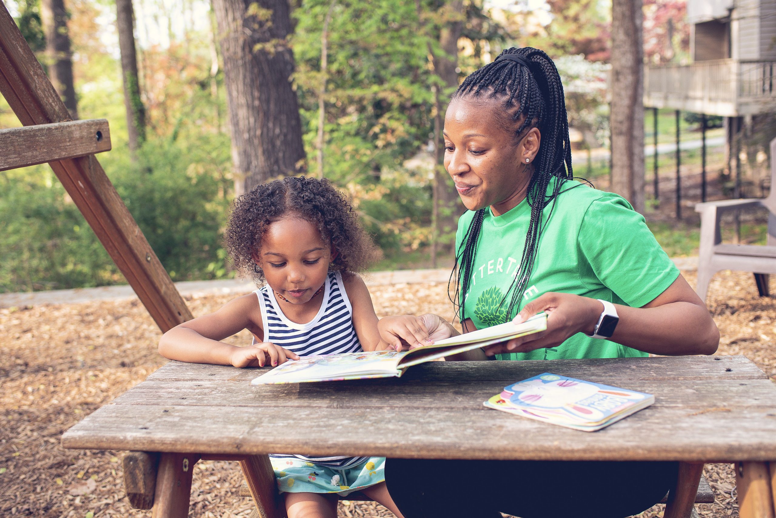 A babysitter wearing a green SitterTree t-shirt sits at an outdoor picnic table with a young girl, reading a colorful book together in a shaded wooded area with a swing set nearby: Nanny cost vs daycare